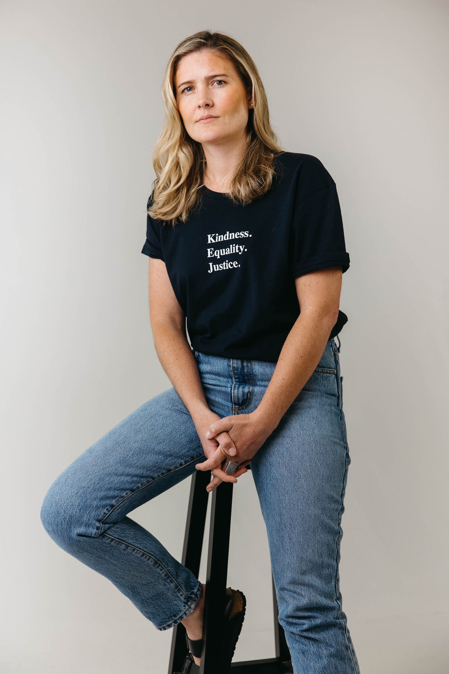 Sophie sitting on a stool smiling wearing an ink blue tshirt with the words 'Kindness Equality Justice.' written in white.