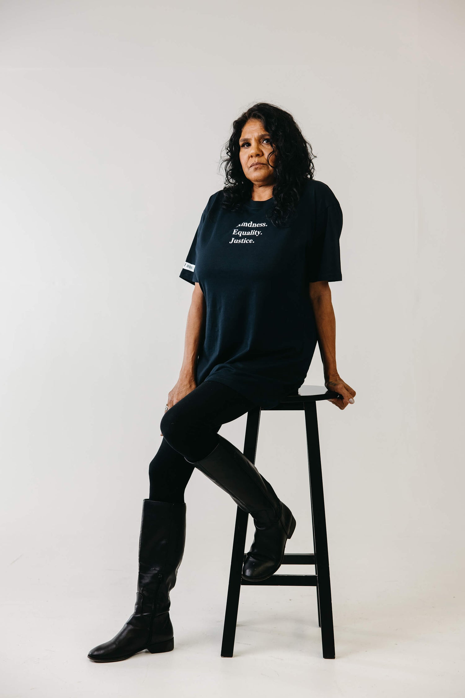 Dell sitting on a stool wearing an ink blue tshirt with the words 'Kindness Equality Justice.' written in white.